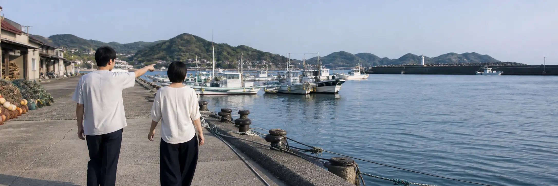 漁港の岸壁を二人が並んで歩き、片方が海側を指している後ろ姿